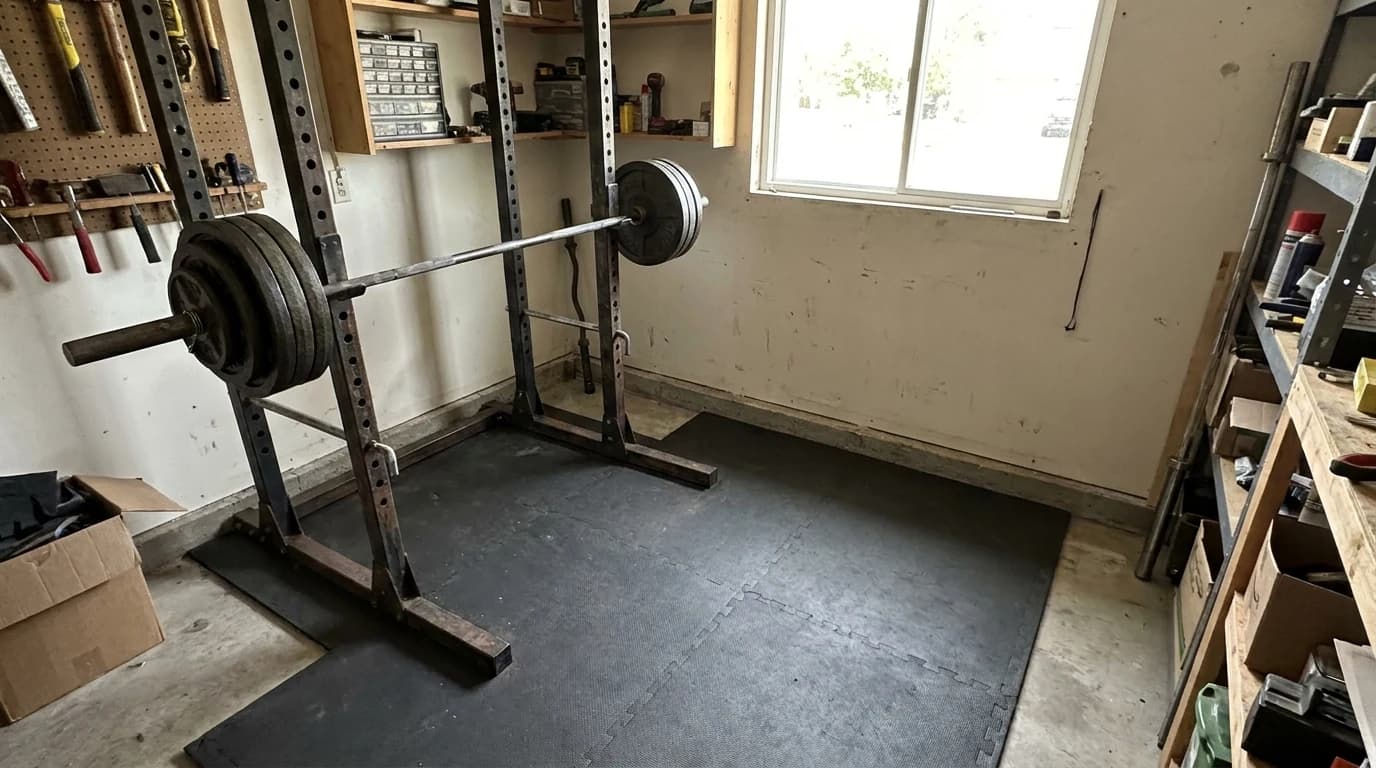 A power rack with a barbell racked in a small garage corner, the bar overhanging the uprights, with clear floor marked out in front of it.