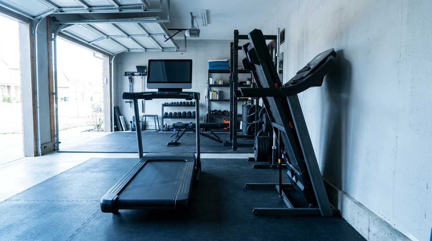 A folding home treadmill with a rotating touchscreen console standing beside a fixed-screen studio treadmill in a garage gym.